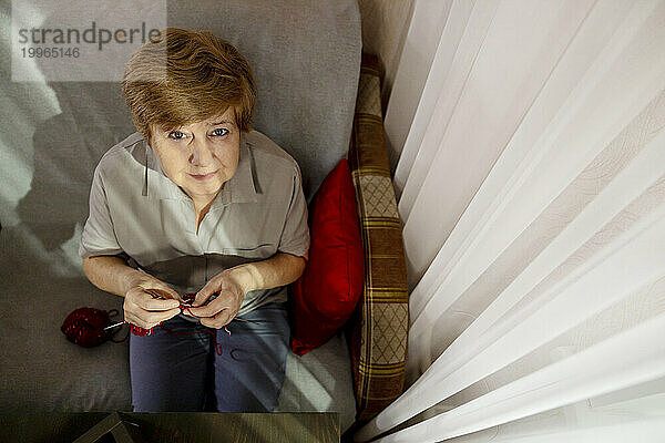 Ältere Frau strickt zu Hause auf Stuhl neben Vorhang