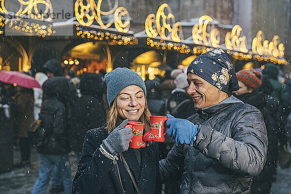 Glückliche Männer und Frauen stoßen auf dem Weihnachtsmarkt auf Tassen Punsch an