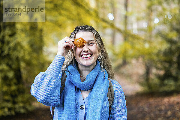 Happy young woman holding leaf over eye in forest