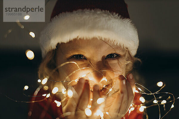 Fröhliches Mädchen mit Weihnachtsmütze und Lichterketten in der Hand