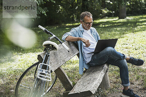 Älterer Mann benutzt Laptop und sitzt mit dem Fahrrad auf Bank im Park