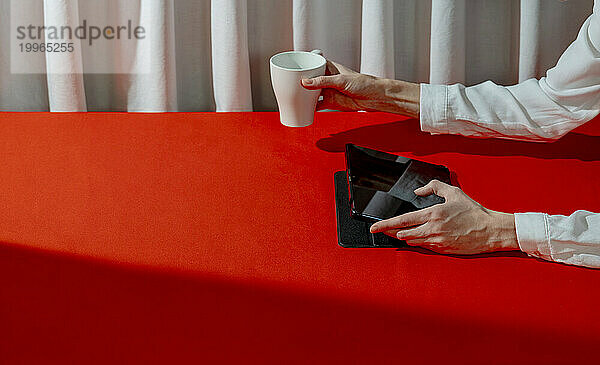 Frau benutzt Tablet-PC und hält eine Tasse Tee am roten Tisch