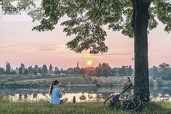 Frau sitzt im Morgengrauen auf Gras unter einem Baum in der Nähe des Flusses