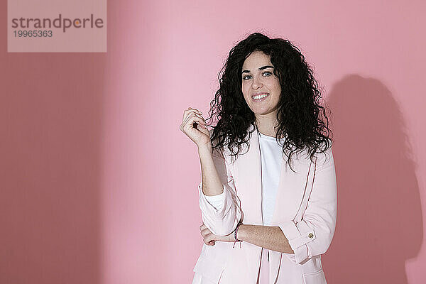 Smiling curly haired woman wearing blazer and standing against pink background