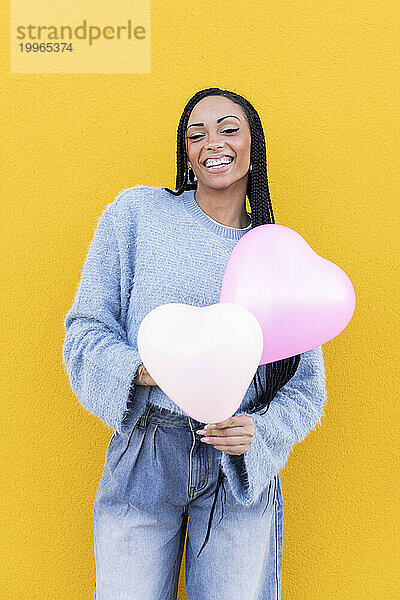 Glückliche junge Frau hält rosa Luftballons vor gelber Wand