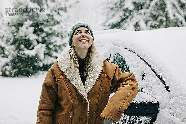 Frau trägt Wintermantel und lehnt im Schnee an Auto