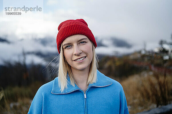 Lächelnde Frau mit roter Strickmütze steht im Winter unter dem Himmel