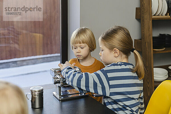 Mädchen und Junge sitzen mit Nudelmaschine am Esstisch