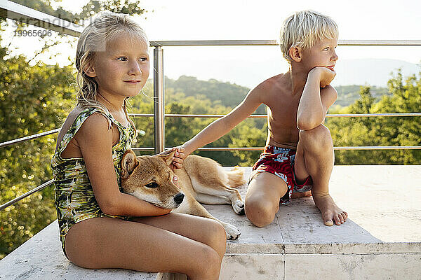 Blondes Mädchen und Junge sitzen an einem sonnigen Tag mit dem Hund Shiba Inu in der Nähe des Geländers