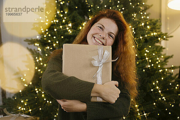 Lächelnde rothaarige Frau hält Geschenkbox neben Weihnachtsbaum