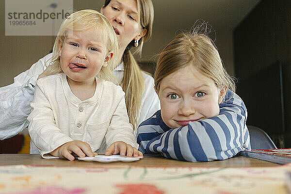 Verspielte Schwestern machen Gesichtsausdrücke mit der Mutter im Hintergrund zu Hause