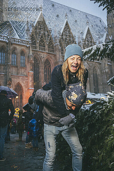 Glücklicher Mann gibt einer Frau Huckepackfahrt auf dem Weihnachtsmarkt