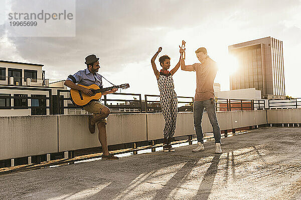 Glücklicher Mann  der Gitarre spielt und mit Freunden auf dem Dach tanzt