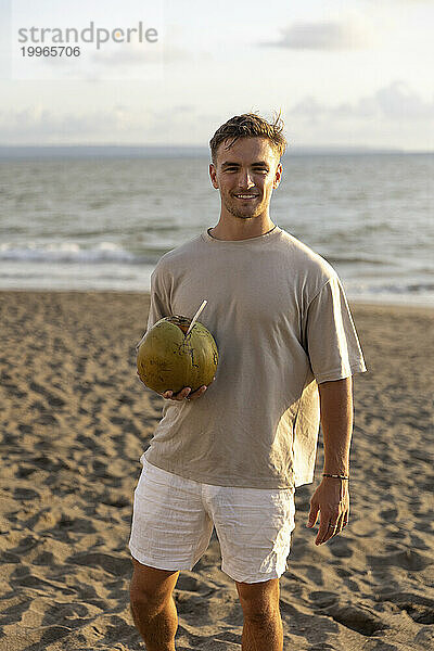 Lächelnder Mann hält frische Kokosnuss bei Sonnenuntergang