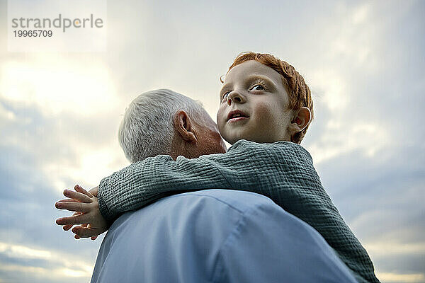Grandfather carrying grandson under cloudy sky