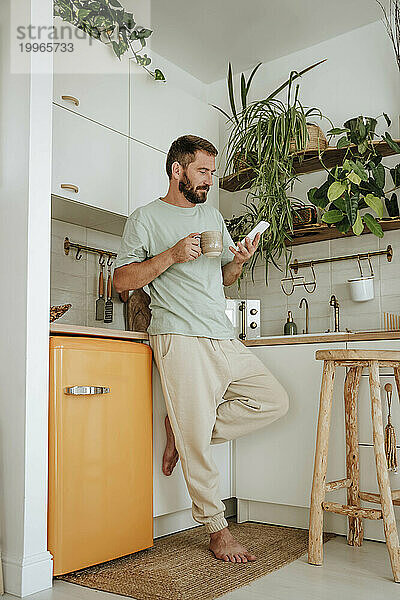 Mann mit Kaffeetasse benutzt Mobiltelefon in der heimischen Küche