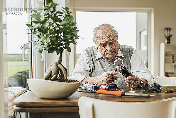 Älterer Mann sitzt am Fenster und untersucht zu Hause den Miniaturzug durch die Lupe