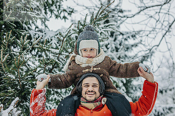 Glücklicher Mann trägt Sohn auf Schultern und hält Händchen in der Nähe eines Baumes