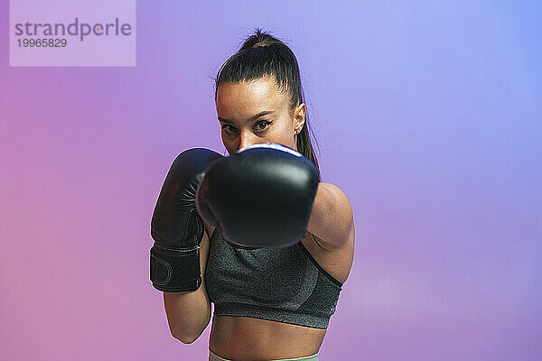 Selbstbewusste junge Frau mit schwarzen Boxhandschuhen trainiert vor farbigem Hintergrund