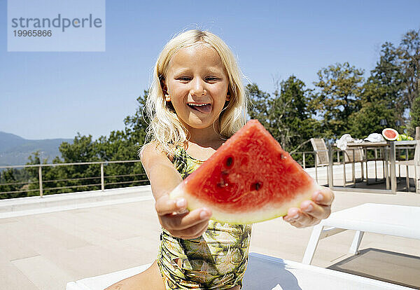 Lächelndes Mädchen  das auf einem Liegestuhl sitzt und eine Scheibe Wassermelone hält