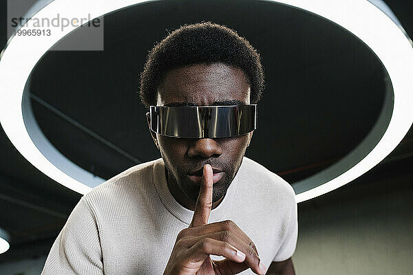Junger Mann mit futuristischer Cyberbrille unter moderner Ringlampe  der den Zeigefinger auf die Lippen legt