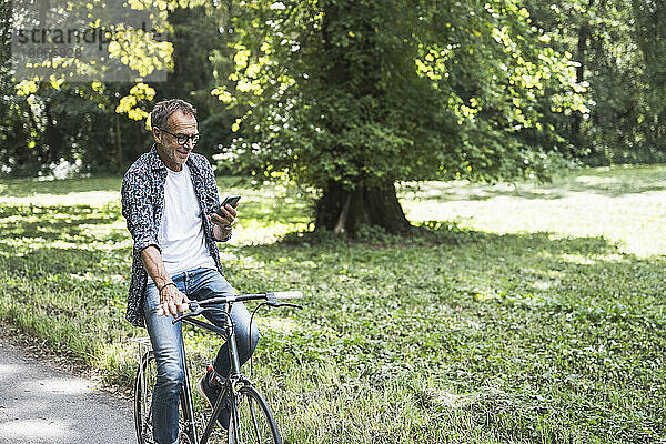 Älterer Mann fährt Fahrrad und benutzt Smartphone im Park