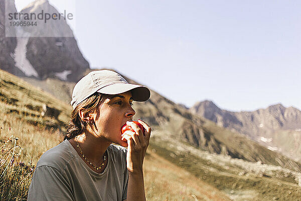 Frau trägt Mütze und isst Tomaten vor den Bergen an einem sonnigen Tag