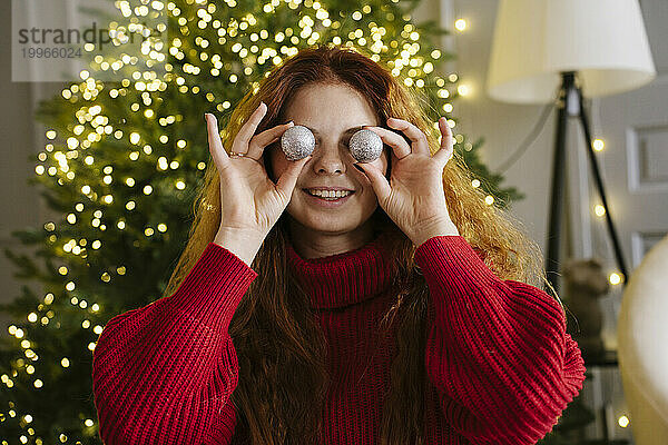 Lächelnde Frau hält zu Hause Weihnachtsschmuck an den Augen