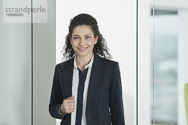 Happy mature businesswoman wearing blazer and standing near glass door in office