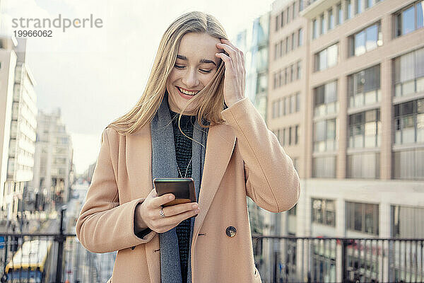 Glückliches blondes Mädchen  das Smartphone vor Gebäuden benutzt