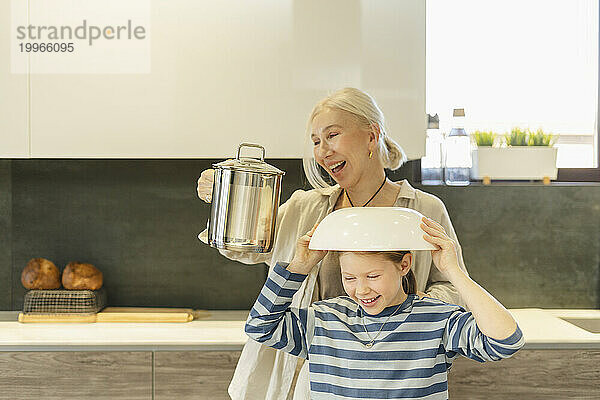 Happy grandmother holding jug and granddaughter with bowl on head enjoying at home