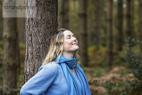 Lächelnde junge Frau steht mit geschlossenen Augen neben einem Baum im Wald