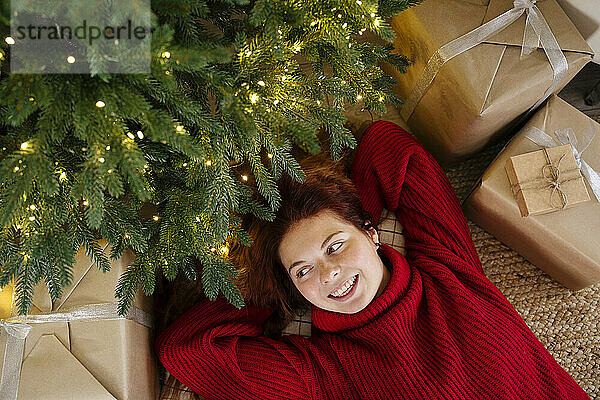 Glückliche Frau  die zu Hause neben Weihnachtsbaum und Geschenkboxen liegt