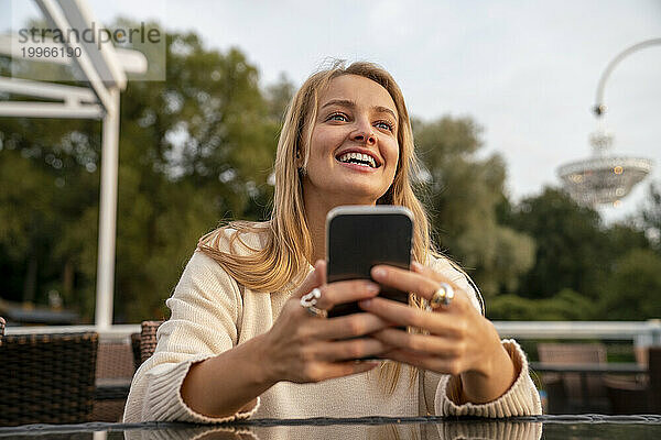 Glückliche blonde Frau sitzt mit Smartphone am Tisch im Café