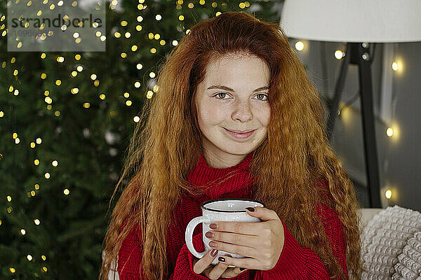 Glückliche rothaarige Frau hält zu Hause eine Kaffeetasse neben dem Weihnachtsbaum