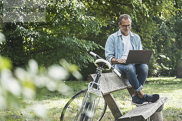 Älterer Mann sitzt mit Laptop auf Bank im Park