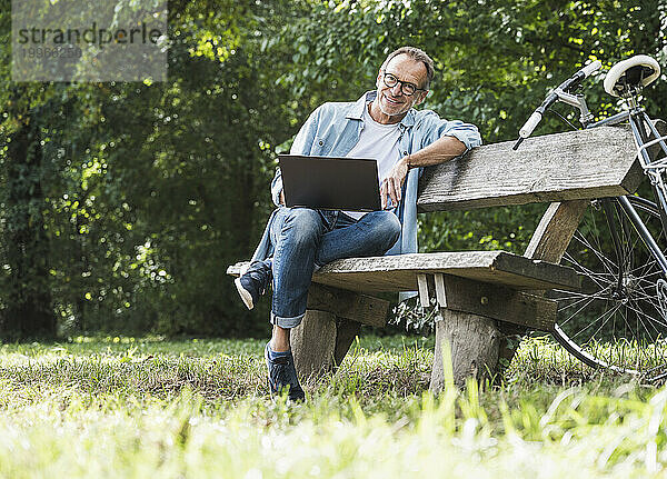 Lächelnder Mann mit Laptop sitzt auf Bank im Park