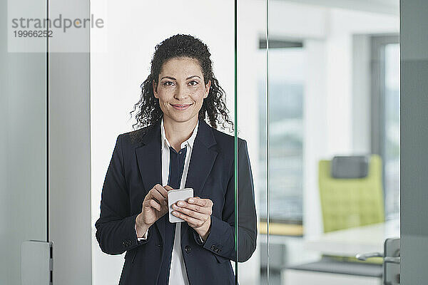 Lächelnde Geschäftsfrau steht mit Mobiltelefon im Büroeingang