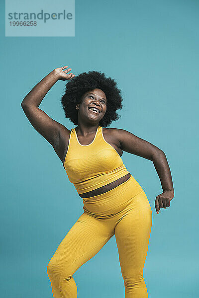 Cheerful young woman in yellow sports clothing dancing against blue background