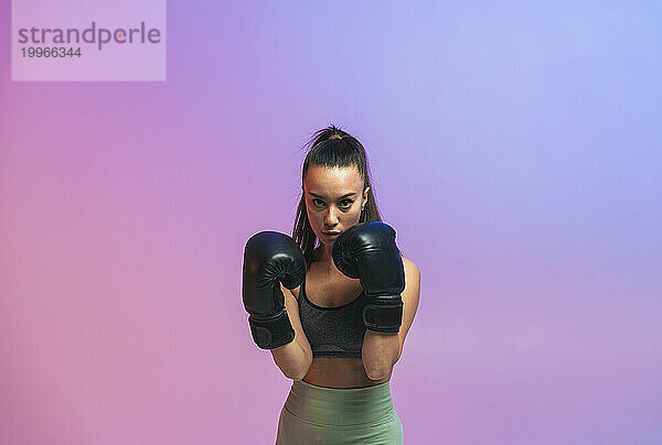 Selbstbewusste junge Frau mit schwarzen Boxhandschuhen vor farbigem Hintergrund