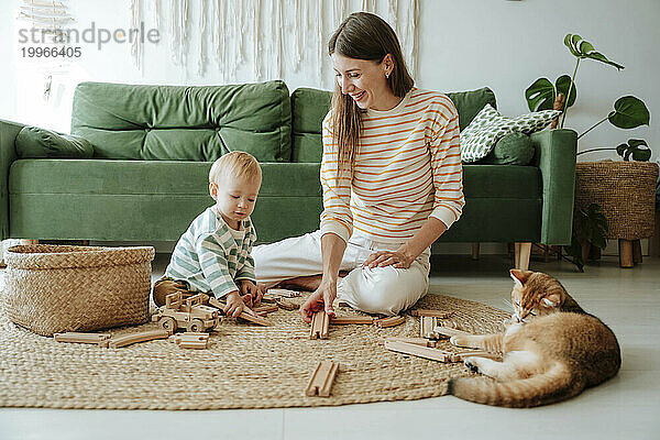 Mutter und Sohn spielen mit einer Holzeisenbahn im Wohnzimmer  beobachtet von einer Katze