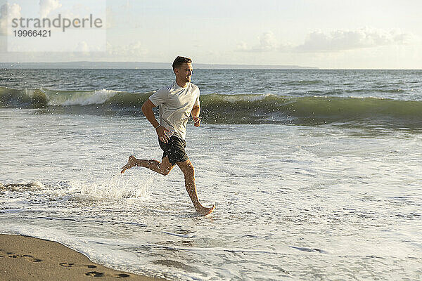 Lächelnder junger Mann  der am Strand im Wasser läuft