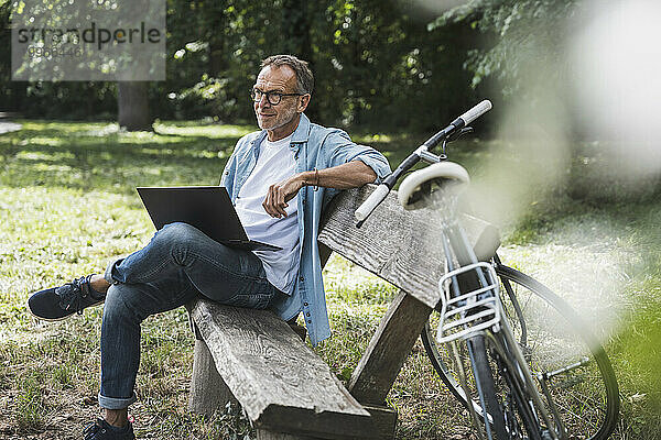 Älterer Mann mit Laptop sitzt auf Bank im Park