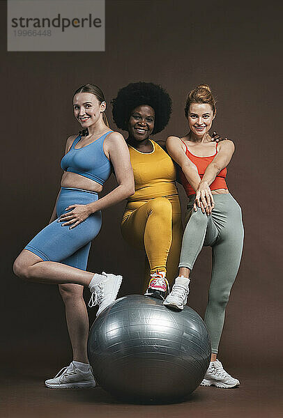 Smiling multiracial friends with fitness ball against brown background