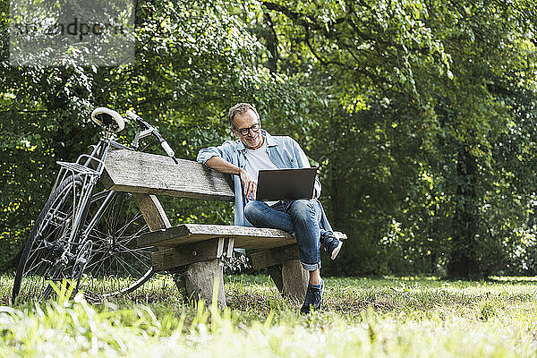 Älterer Mann sitzt am Wochenende mit Laptop auf Bank im Park