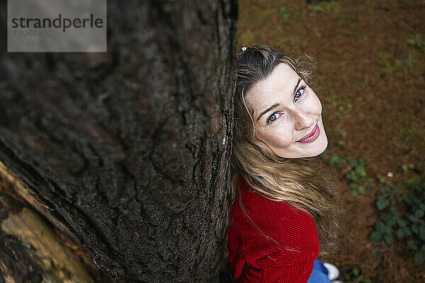 Lächelnde schöne Frau lehnt an Baumstamm im Wald