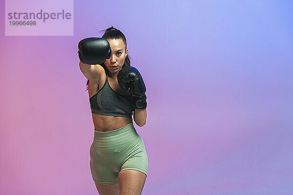 Selbstbewusste junge Frau mit schwarzen Boxhandschuhen übt vor farbigem Hintergrund