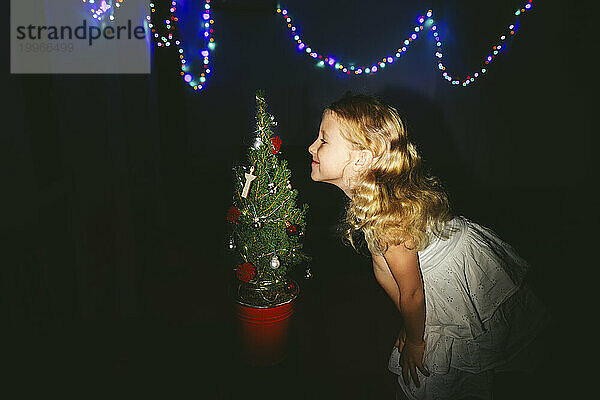 Lächelndes Mädchen riecht geschmückten Weihnachtsbaum in der Nähe von Lichterketten