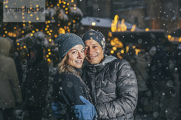 Lächelndes Paar steht im Schnee auf dem Weihnachtsmarkt