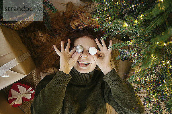 Smiling woman holding Christmas ornaments on eyes at home